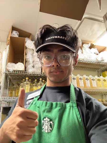 Salman giving a thumbs up in a green Starbucks apron and visor in front of syrup shelves.